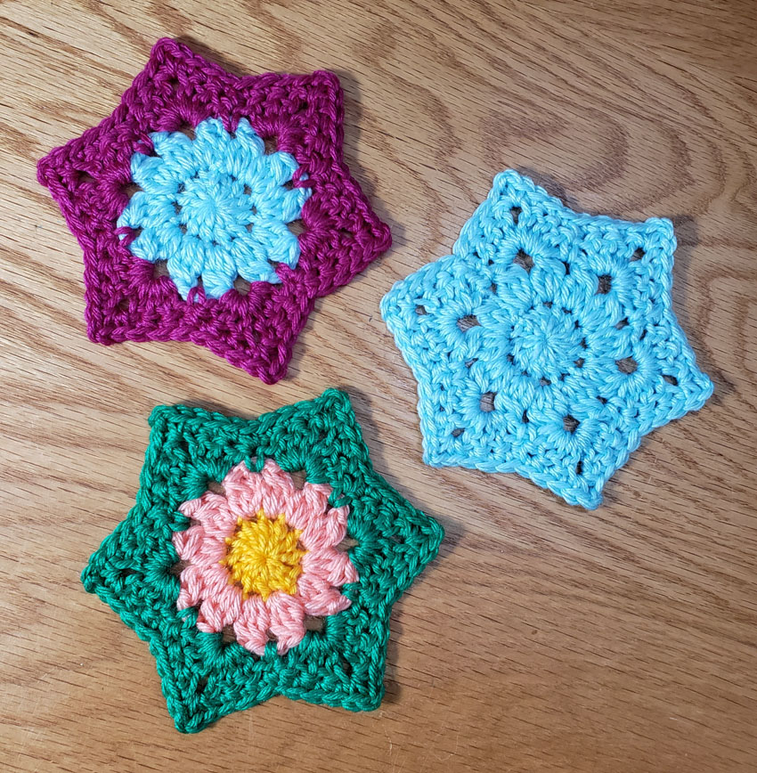 3 hexagonal crocheted motifs, 1 in single color of light blue, 1 in 2 colors of light blue in center with violet outside, 1 with yellow center surrounded by peach colored petals finished with bright green as leaves.