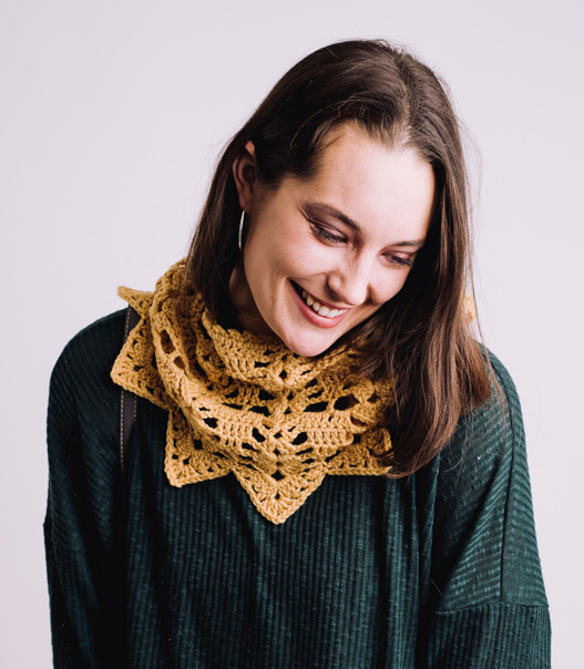 Caucasian model with should length brown hair and a dark green long sleeve shirt looking down Around her neck is golden yellow lacy cowl with points along the bottom edge.
