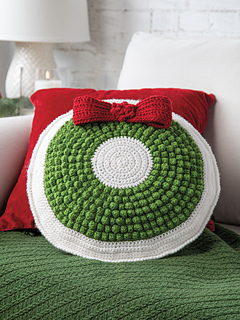 Background is a white brick wall with a white lap and candle in left corner. Foreground is a white sofa with a red square pillow sitting on top of a green textured blanket on the seat of sofa. In center of image and in front of red pillow is a round white pillow with a textured round bright green circle topped by a a red 3d bow creating a Christmas wreath on the pillow.