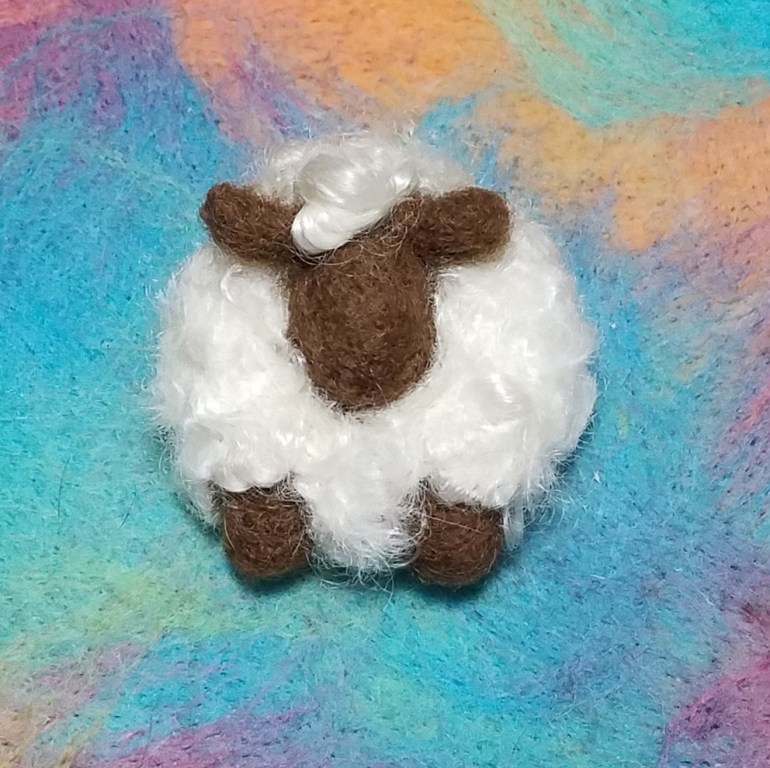 An image of the pin which is a little round base relief sheep front view with curly white fleece, simple brown face and ears and little round brown front legs on an abstract colorful background.
