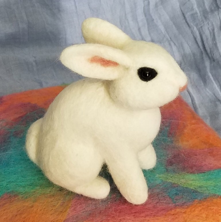 Main image is a photograph of a 3 dimensional white bunny sitting on it's back legs. Inside bunny's ear is pink with a pink nose and dark black shiny eye. Bunny is sitting on a multi-colored surface with a gray background.